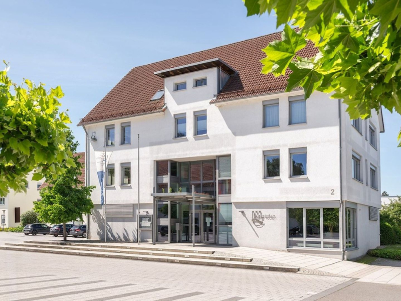 Ein modernes, dreistöckiges weißes Gebäude mit einem roten Dach, umgeben von grünen Bäumen und einem gepflasterten Vorplatz.