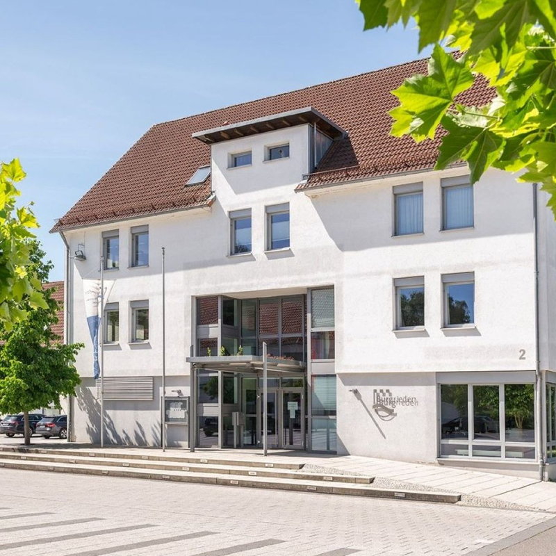 Ein modernes, dreistöckiges weißes Gebäude mit einem roten Dach, umgeben von grünen Bäumen und einem gepflasterten Vorplatz.