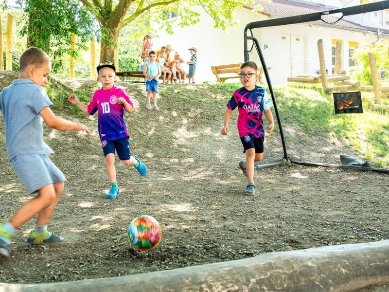 Mehrere Kinder spielen Fußball im Freien auf einem kleinen Hügel unter Bäumen.