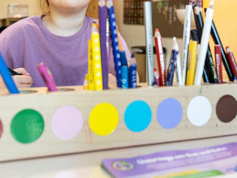 Ein fröhliches Kind sitzt an einem Tisch in einem farbenfrohen Klassenzimmer, umgeben von Stiften und Malutensilien, während es zeichnet, und weitere Kinder sind im Hintergrund zu sehen.