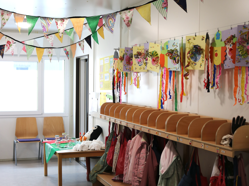 Ein bunter Kindergartenflur mit hängenden Wimpelgirlanden, Jacken und künstlerischen Wandarbeiten, sowie einer Anrichte mit Spielzeug und Stühlen.