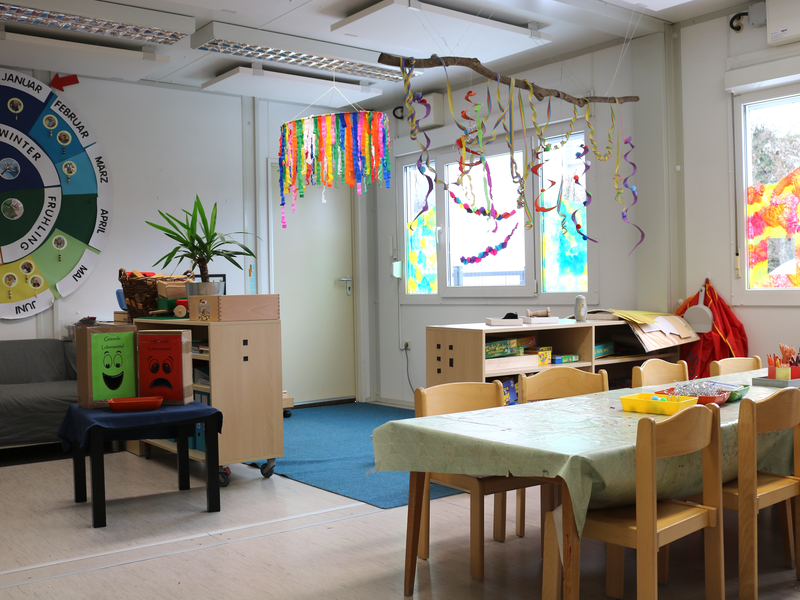 Ein bunter und einladender Kindergartenraum mit einem Tisch und Stühlen, dekoriert mit farbenfrohen Hängedekorationen, einem Jahreszeitenrad, Pflanzen und kreativen Bastelmaterialien.