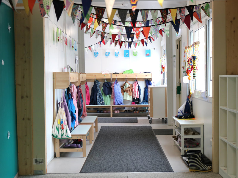 Ein farbenfroher Kindergartenflur mit an der Decke aufgehängten Wimpeln, an den Seiten bunte Jacken und Taschen an Garderobenhaken und Regale für Schuhe.