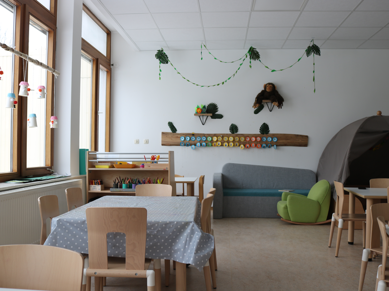 Ein heller Kindergartenraum mit Holzmöbeln, einem Spielzelt, hängenden Basteleien vor den Fenstern, Wanddekorationen und farbenfrohen Sitzmöglichkeiten.