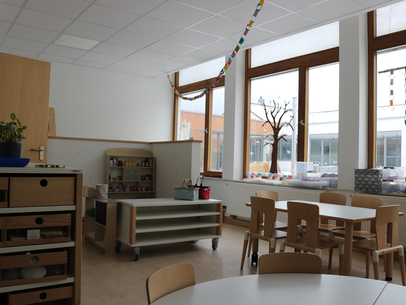 Ein heller Raum mit mehreren Tischen und Stühlen, großen Fenstern, einem Regal mit Spielzeugerkennbar und bunten Papierketten an der Decke.