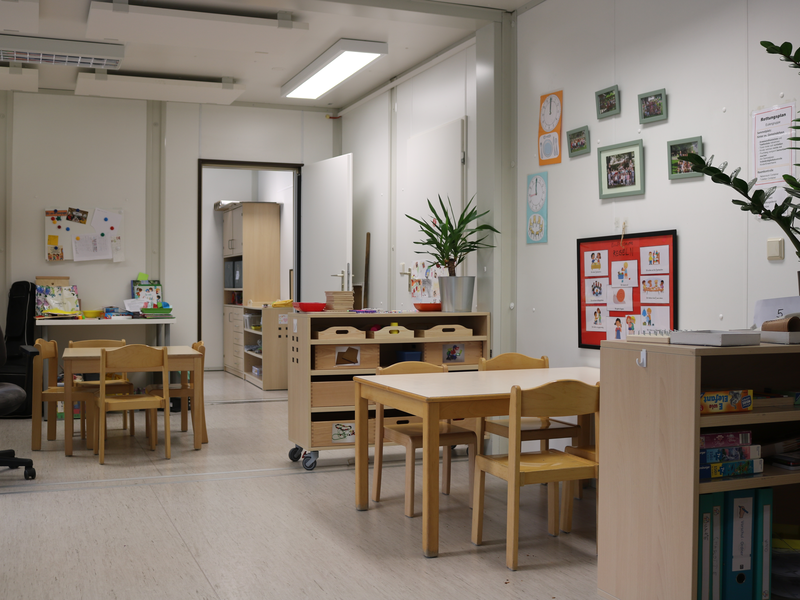 Das Bild zeigt einen hellen, freundlich eingerichteten Kindergartenraum mit Tischen, Stühlen, Pflanzen und bunten Zeichnungen an den Wänden.