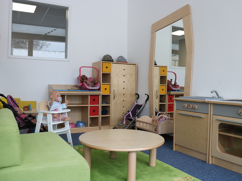 Ein Kinderzimmer mit Spielküche, Puppenwagen, Spiegel und einem Tisch samt Sofa, das mit Spielzeug und Puppen bestückt ist.