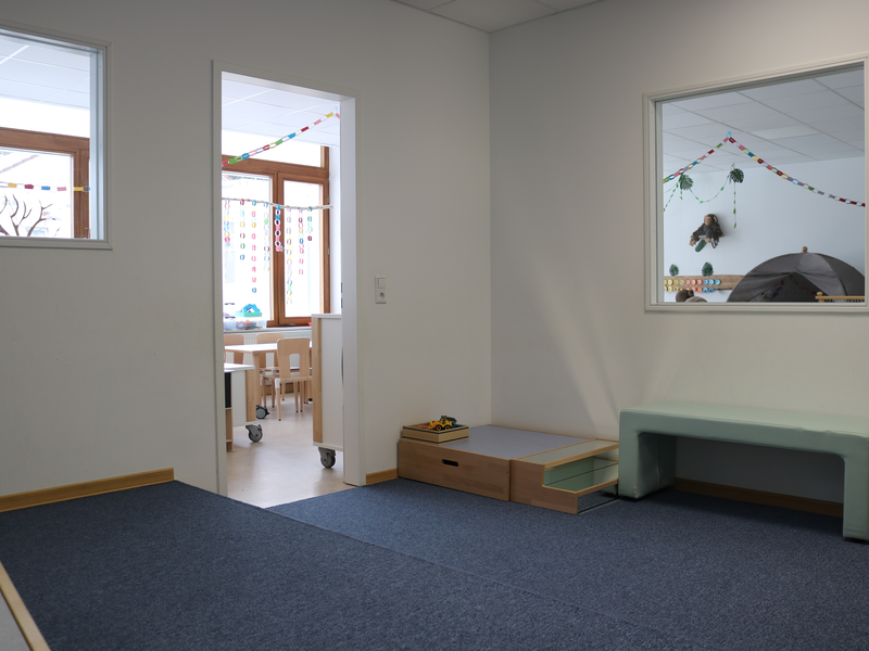Ein heller Nebenraum mit blauer Auslegware zeigt einen Durchgang zu einem helleren Kindergartenraum, teilweise sichtbar sind bunte Papiergirlanden und Kindermöbel.