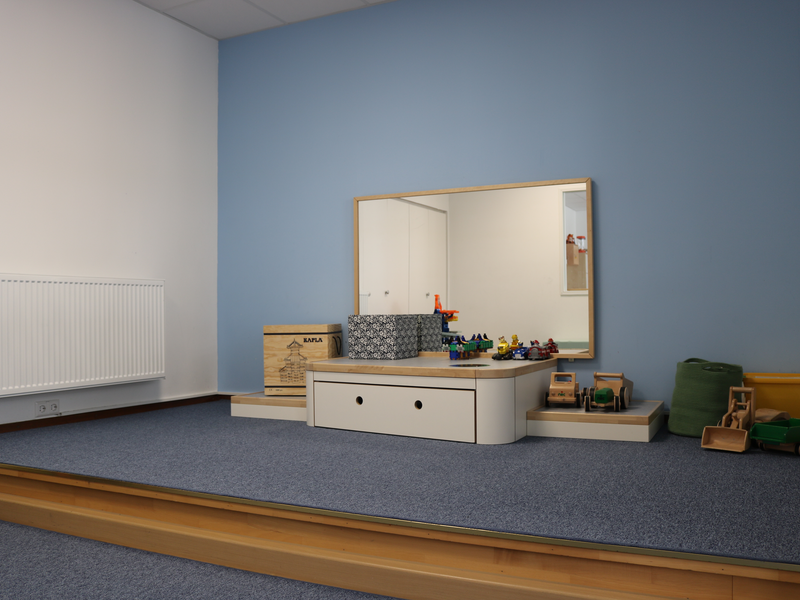 Ein schlicht eingerichteter Raum mit einer erhöhten Plattform, auf der ein großer Spiegel, Holzspielzeug und Kisten platziert sind.
