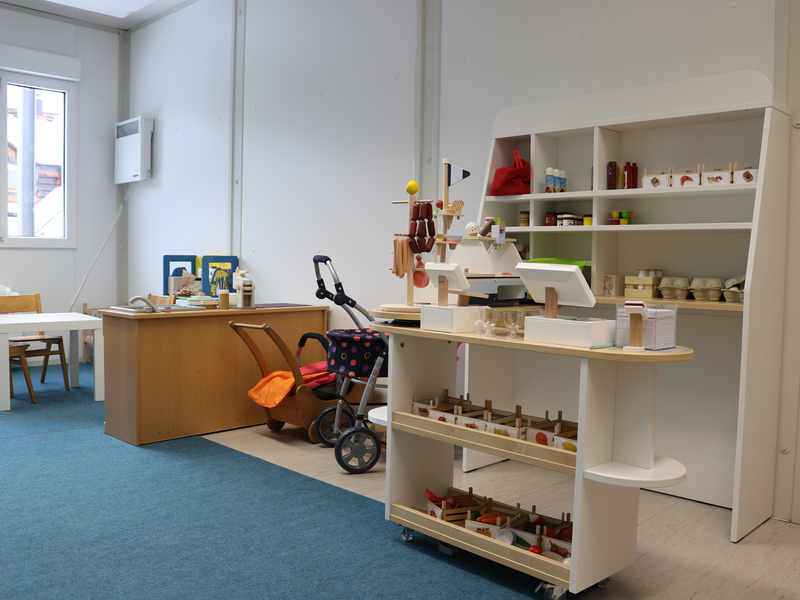 Ein heller Raum mit einem kleinen Tisch und Stühlen, einem Holzspielzeuggeschäft und einem Kinderwagen.