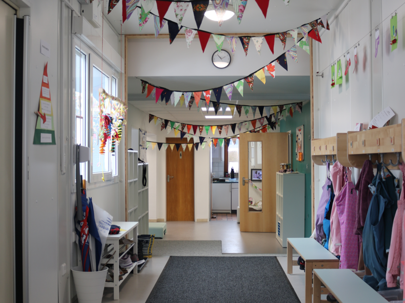 Ein Flur in einer Kita mit bunten Wimpelgirlanden an der Decke, Kinderjacken an Haken und Regalen für Schuhe.