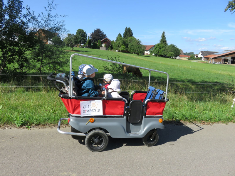 Ein modernes Transportgefährt mit zwei Kindern darin fährt entlang einer ländlichen Straße mit grünen Wiesen im Hintergrund.
