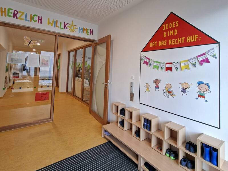 Ein Eingangsbereich einer Kita mit einem Schild "Herzlich Willkommen", Schuhregalen und einer farbenfrohen Wanddekoration, die auf die Rechte der Kinder hinweist.