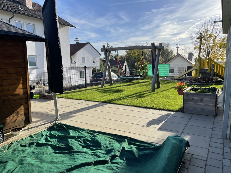 Ein sonniger Außenbereich einer Kinderkrippe mit einer Schaukel, einer Rutsche, Rasenfläche und Pflanzkästen.