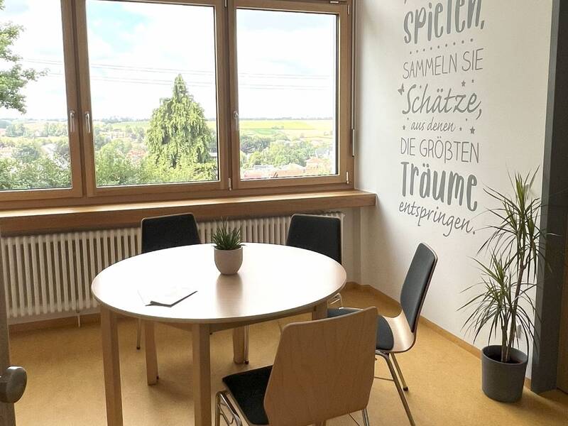 Ein heller Raum mit einem runden Tisch und vier Stühlen, dekoriert mit einer Wandinschrift und einem Pflanztopf auf dem Tisch.