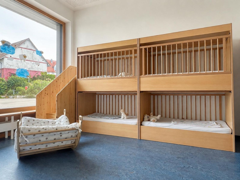 Ein heller Raum mit vier Holzgitterbetten, einem großen Fenster und einem kleinen Tisch mit gepolsterten Körbchen, auf einem blauen Boden.