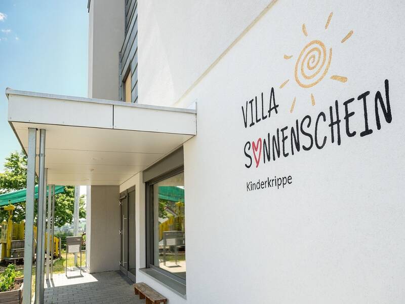 Außenansicht eines Gebäudes mit der Aufschrift "Villa Sonnenschein Kinderkrippe" und einem gemalten Sonnensymbol.