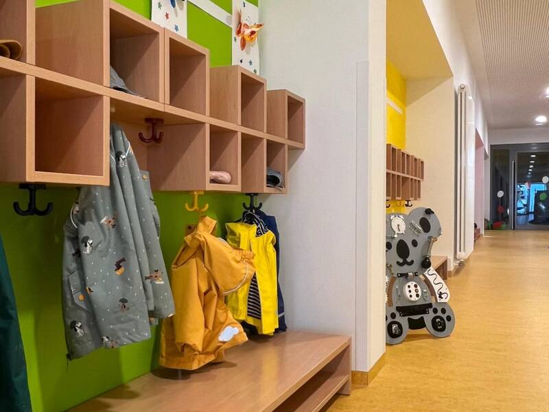 Ein bunter Kindergarten-Garderobenbereich mit bunten Jacken, offenen Holzfächern und kindgerechten Wanddekorationen.
