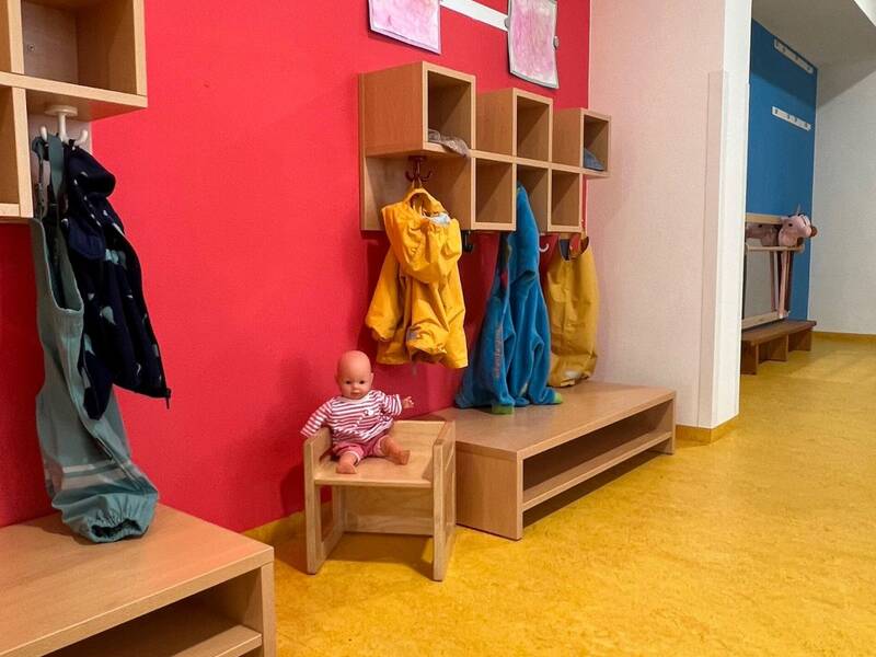Ein Kindergartenflur mit einer roten Wand, an der Garderobenhaken mit Jacken und ein kleines Holzregal angebracht sind, sowie eine Puppe, die auf einem Holzstuhl sitzt.
