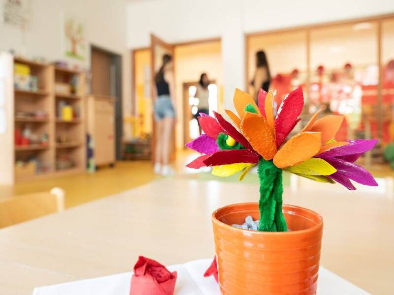 In einem Raum steht eine bunte, aus Papier gebastelte Blume in einem orangefarbenen Topf im Vordergrund, während Menschen im Hintergrund unscharf abgebildet sind.
