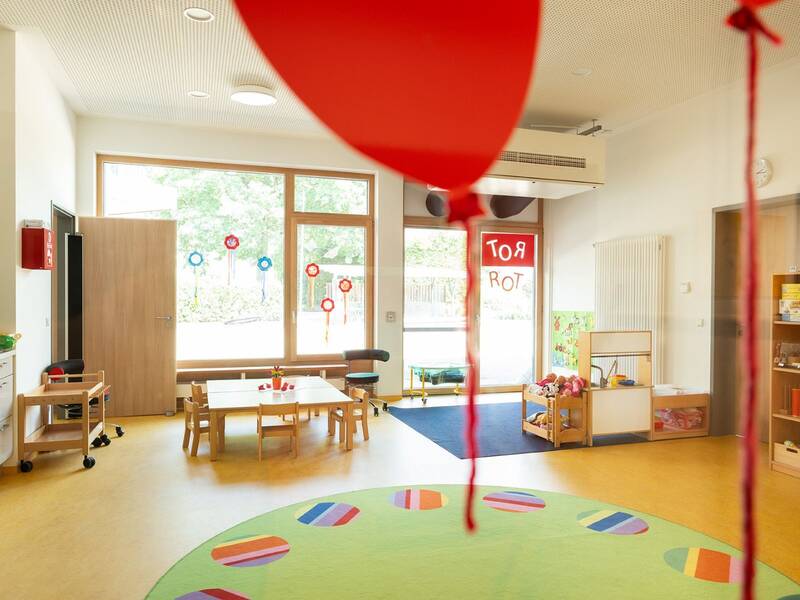 Ein bunter Krippenraum mit großen Fenstern, Tischen, Stühlen, Spielzeug und roten Luftballons im Vordergrund.