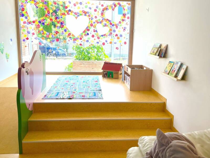 Der Raum zeigt einen hellen Spielbereich mit bunten Fensterdekorationen, einer Spielzeug-Ecke und einem großen Plüschelefanten auf einem Sitzsack.