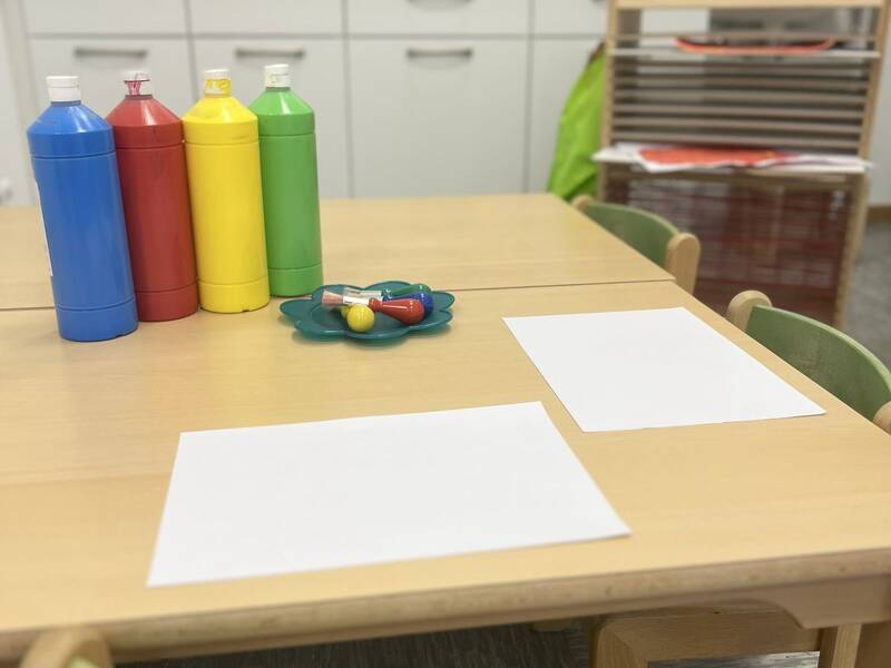Ein Tisch in einem Raum, auf dem vier bunte Flaschen und zwei leere Blätter Papier liegen, mit einer Küche im Hintergrund.