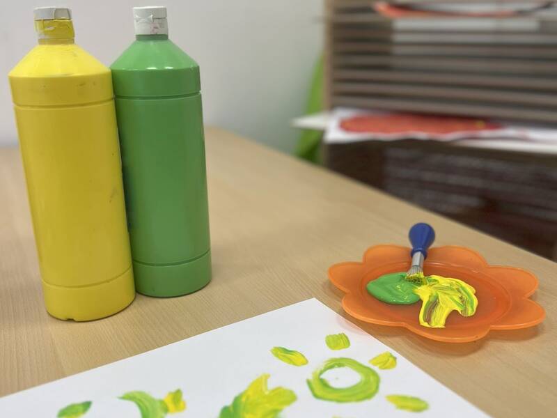 Auf einem Tisch stehen zwei Farbflaschen in Gelb und Grün sowie ein orangefarbener Teller mit einem Pinsel, daneben befindet sich ein Blatt Papier mit einem Fingerbild in den gleichen Farben.