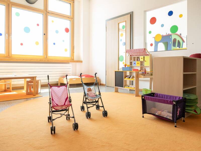 Heller Raum in einer Kindertagesstätte mit bunten Punkten an den Fenstern und Spielzeugkinderwagen auf einem orangefarbenen Teppich.