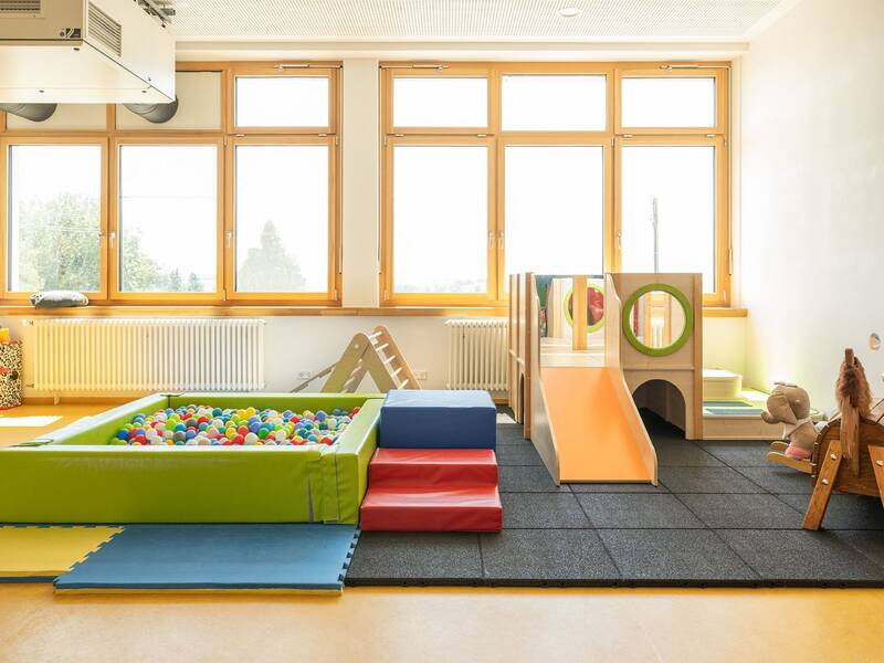 Ein heller Spielraum mit großen Fenstern zeigt einen Kletterbereich mit Rutsche und ein Bällebad.