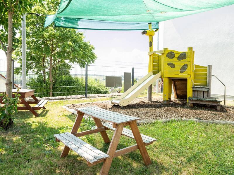 Ein Spielplatz mit einem überdachten Bereich, einer Holzrutsche, die wie eine Giraffe aussieht, und einer Picknickbank unter Bäumen.