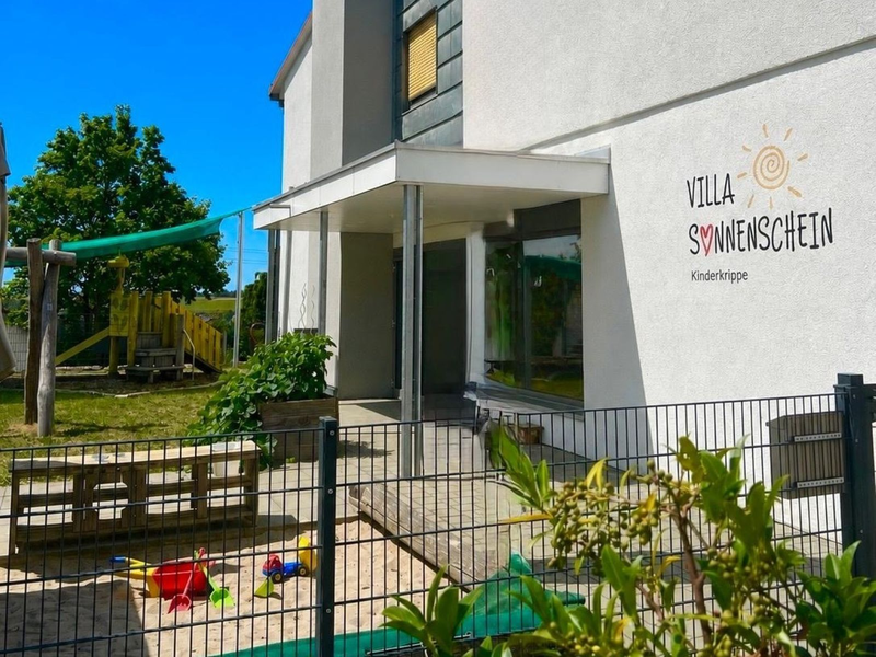 Das Bild zeigt das Gebäude einer Kinderkrippe namens "Villa Sonnenschein" mit einem Spielplatz im Freien, bestehend aus Rutsche, Sandkasten und buntem Spielzeug.