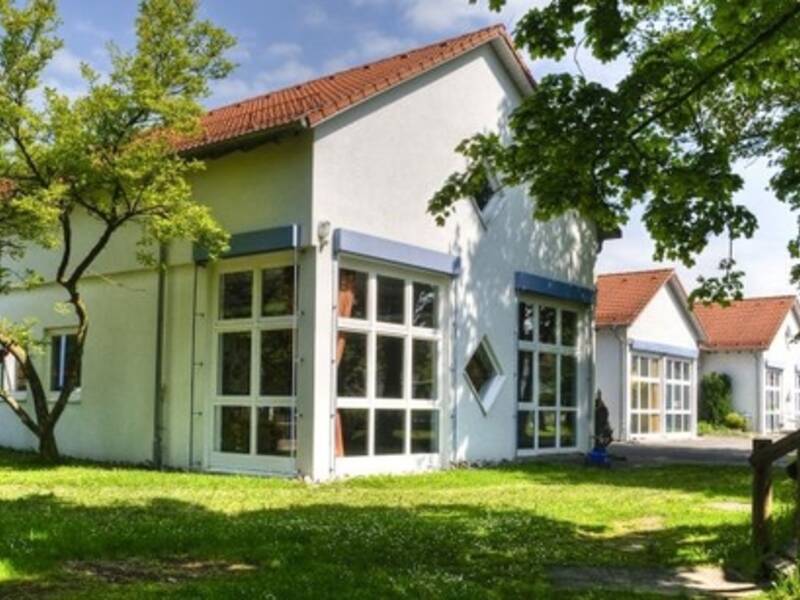 Das Bild zeigt mehrere weiße Gebäude mit roten Dächern, die von grüner Vegetation umgeben sind. Es handelt sich um das Gebäude des Kindergartens.