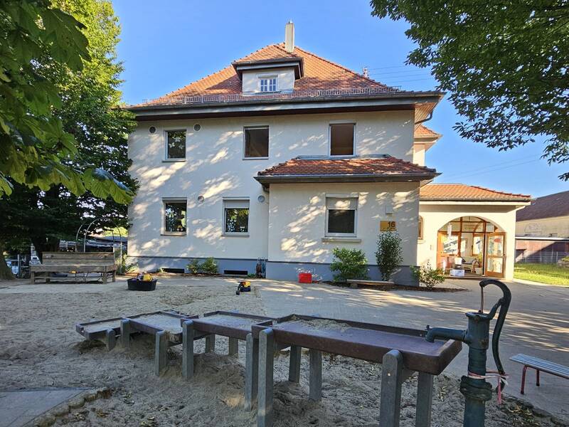 Ein helles, zweistöckiges Gebäude mit rotem Dach steht bei Sonnenschein inmitten von Bäumen, umgeben von einem Spielplatz mit Sand und Spielgeräten.