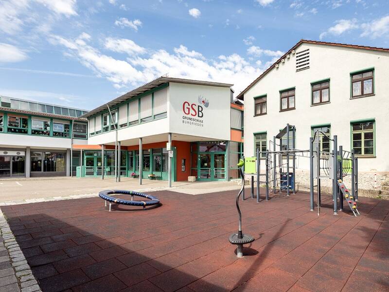 Der Schulhof einer Grundschule zeigt ein Klettergerüst und eine Wippe im Vordergrund, mit einem modernen Schulgebäude im Hintergrund.