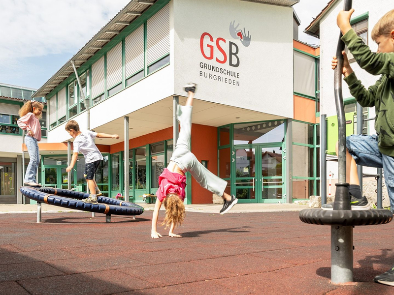 Einige Kinder spielen auf einem Schulhof vor einem Gebäude mit der Aufschrift "GSB Grundschule Burgrieden".