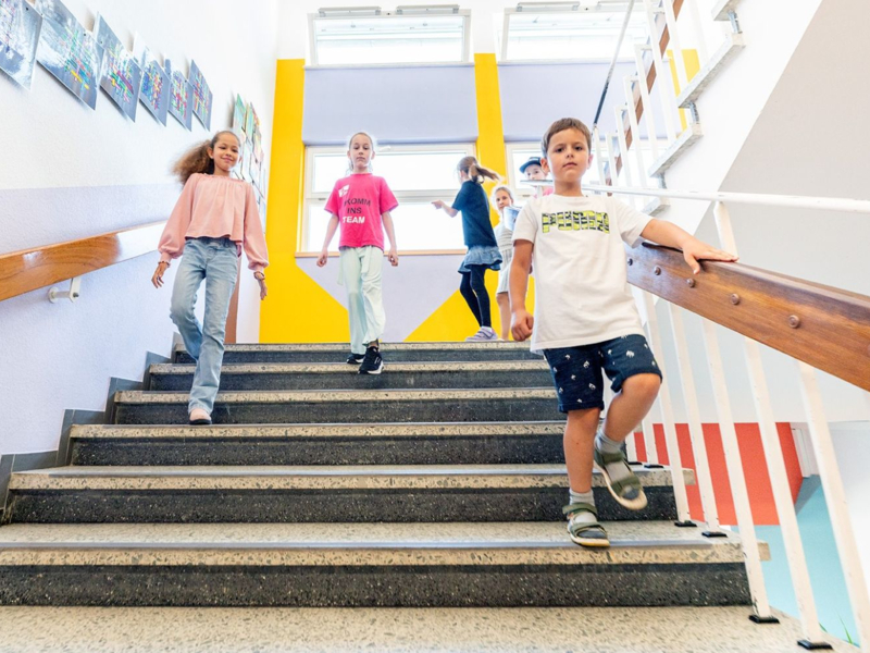 Mehrere Kinder gehen eine Treppe in einem Schulgebäude hinunter, an den Wänden hängen bunte Kunstwerke.