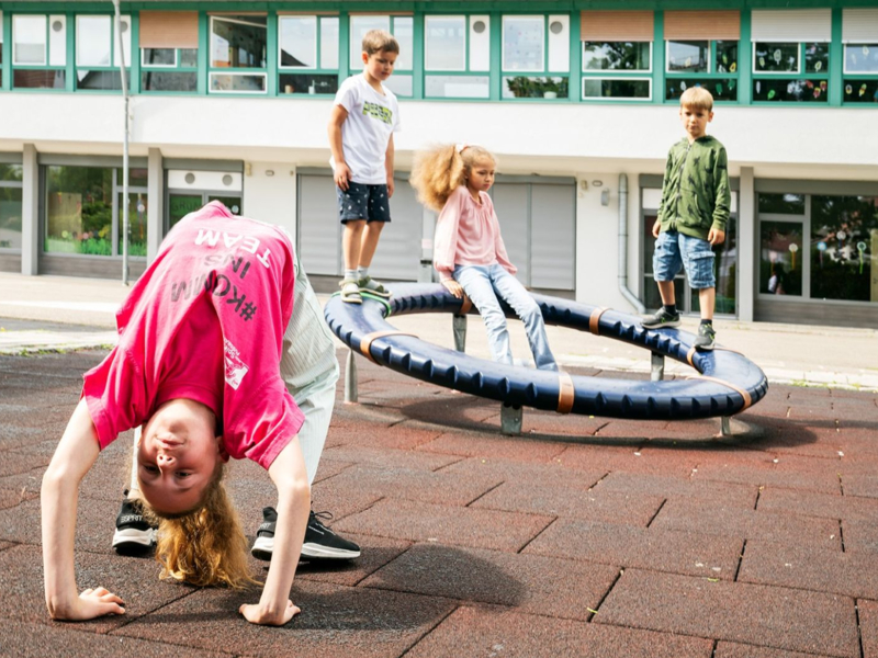 Ein Kind macht einen Handstand auf einem Schulhof, während drei andere Kinder auf einem kreisförmigen Klettergerät balancieren.