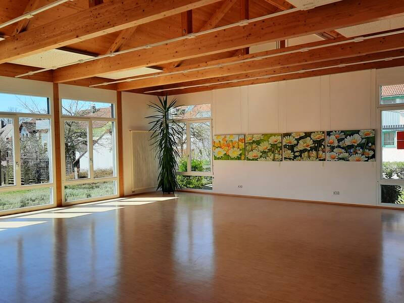 Ein heller Raum mit großen Fenstern, Holzbalkendecke und mehreren Bildern an der Wand.